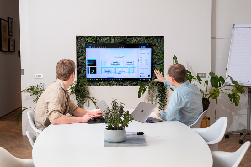 Twee teamleden bekijken samen een website-interface op een groot scherm.